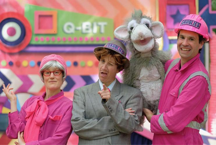 Three performers posing in pink outfits with a puppet rat